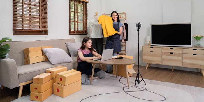 Lesbian couple managing an online live selling business from home, showcasing clothing products and preparing shipping boxes.