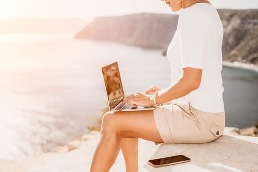 Digital nomad, Business woman working on laptop by the sea. Pretty lady typing on computer by the sea at sunset, makes a business transaction online from a distance. Freelance remote work on vacation