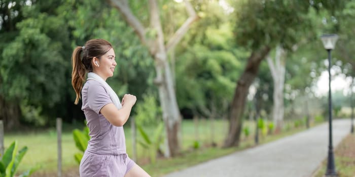 A woman running in a park during the morning, wearing earphones and a towel around her neck, enjoying her exercise routine amidst nature.