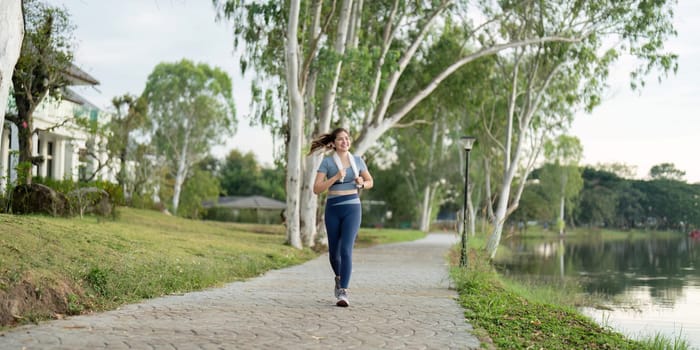 A woman jogging along a lakeside path in a park during the morning, surrounded by trees and greenery, promoting a healthy and active lifestyle.