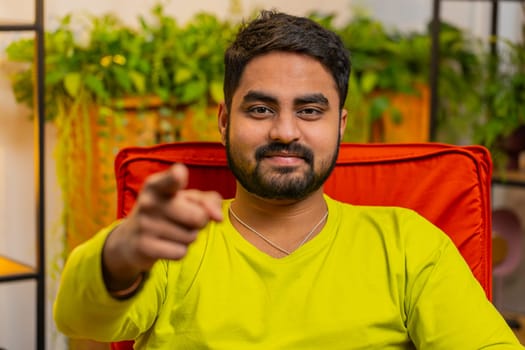 Hey you. Happy satisfied Indian man smiling excitedly and pointing to camera, choosing lucky winner, indicating to awesome you, inviting, approve. Arabian Hindu guy at home room sitting on chair