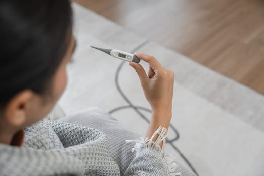 A woman wrapped in a blanket checks her temperature with a digital thermometer, indicating she is sick with a fever.