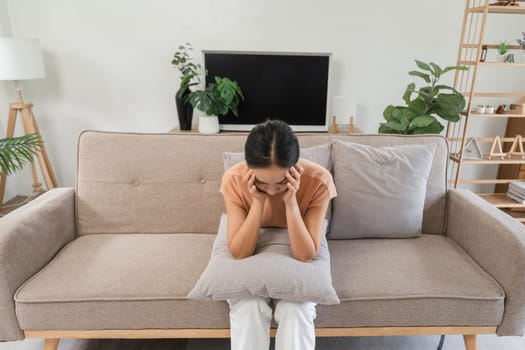 Young woman sitting on a sofa, feeling sick with a headache, holding a pillow for comfort, expressing discomfort and pain in a home environment.