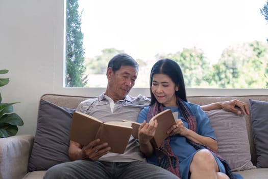 An elderly couple enjoys reading books together on a comfortable sofa in a well lit living room, sharing a peaceful moment.