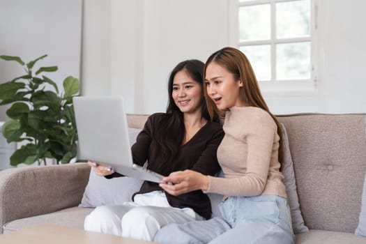 Two smiling lesbian women sitting on a couch, using a laptop together in a bright, modern living room.