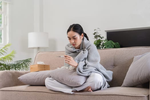 Young woman feeling unwell, wrapped in a blanket, checking her temperature with a thermometer while sitting on a couch at home.