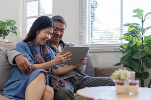 An elderly couple sitting together on a couch, smiling and using a tablet in a bright, cozy living room with plants and natural light.