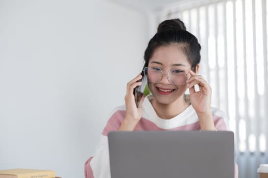 A young woman studies online using her laptop, smiling while talking on the phone, in a bright and modern home office setting.