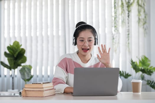 A young woman wearing headphones studies online using a laptop at home. She is surrounded by books and a coffee cup in a bright, modern room with plants.