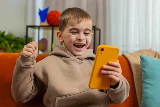 Happy child boy use mobile smartphone typing playing browsing found out great big surprise, good positive news celebrate online game win. Amazed teenager little kid at home in living room sits on sofa