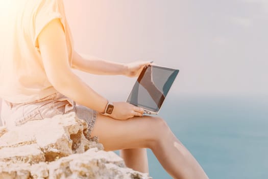 Digital nomad, woman in the hat, a business woman with a laptop sits on the rocks by the sea during sunset, makes a business transaction online from a distance. Freelance, remote work on vacation.