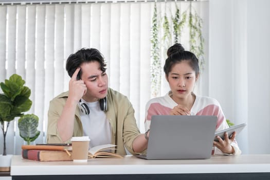 Two students study together online using a laptop and tablet, surrounded by books and coffee in a modern home environment.