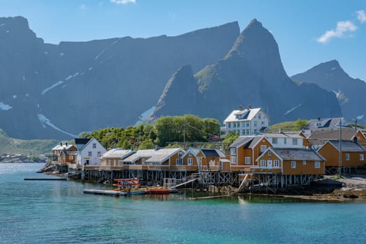 A quaint village of colorful houses on stilts sits on the edge of a calm fjord in Norway, with towering mountains rising in the background. Yellow fisherman Rorbuer cabins , Sakrisoy,Lofoten, Norway