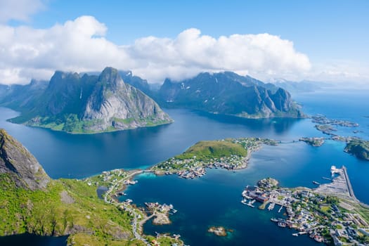 A breathtaking aerial view of a charming village nestled in a stunning Norwegian fjord. Rolling green hills and majestic mountains surround the clear blue waters. Reinebringen, Lofoten, Norway