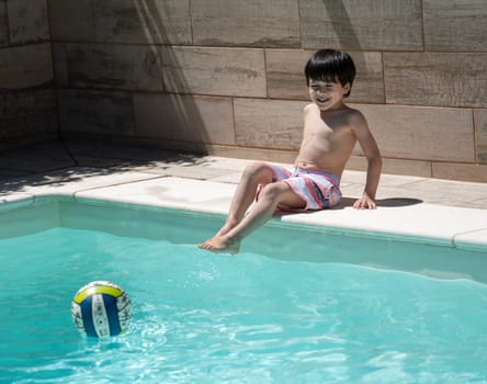 Dominican young boy enjoys the pool on holidays sitting and looking at his ball toy.