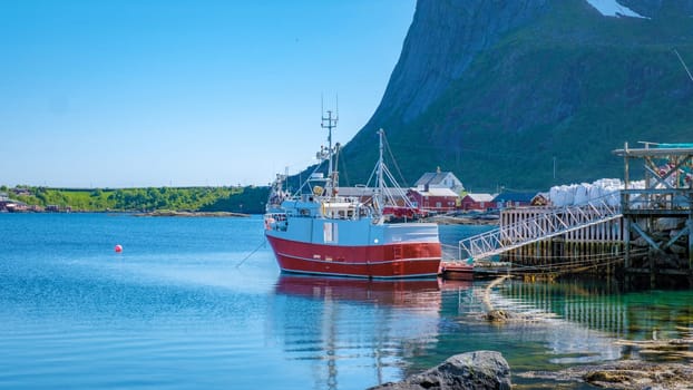 A red and white fishing boat is docked at a pier in a small Norwegian village. The boat is surrounded by calm water and a mountain in the background. Reine, Lofoten, Norway