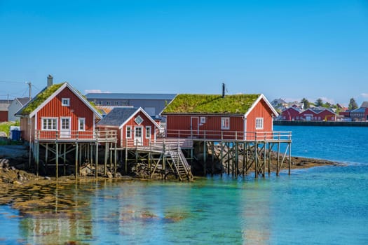 Three red cabins on stilts stand on a rocky outcropping in a Norwegian fjord, with turquoise water reflecting the clear blue sky. Reine, Lofoten, Norway