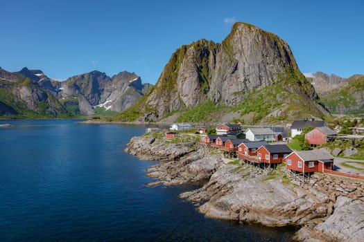 red cabins perched on the rocky shore of a fjord in Norway, surrounded by towering mountains and a clear blue sky. Hamnoy fishing village on Lofoten Islands, Norway with red rorbu houses