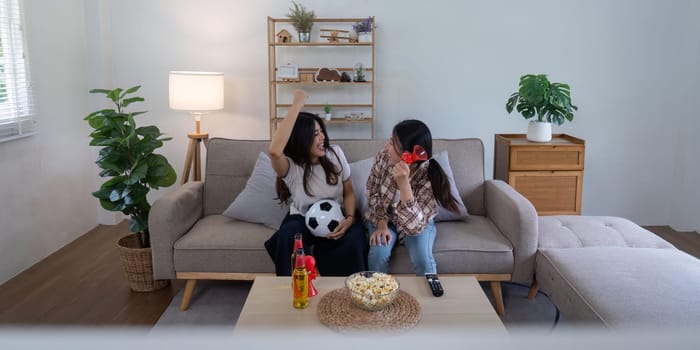 Lesbian couple celebrating a football victory together on a cozy sofa, surrounded by snacks and drinks in a modern living room.