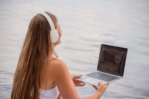 A woman is sitting by the water with a laptop in her lap. She is wearing headphones and she is listening to music. Concept of relaxation and leisure