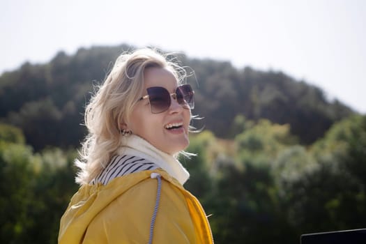 A woman wearing a yellow jacket and sunglasses is smiling. She is standing in a forest with trees in the background