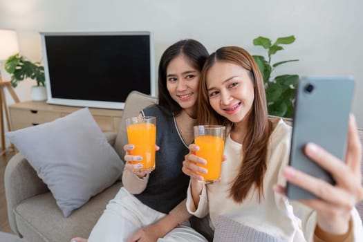 Smiling lesbian couple sitting on a couch, drinking fresh orange juice, and taking a selfie in their cozy home.