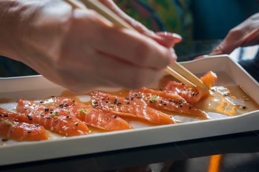 A woman with chopsticks in hand takes thin slices of salmon marinated with sesame, herbs and sauce. Healthy vegetarian food, raw salmon meat. Delicious Asian dish served on restaurant table