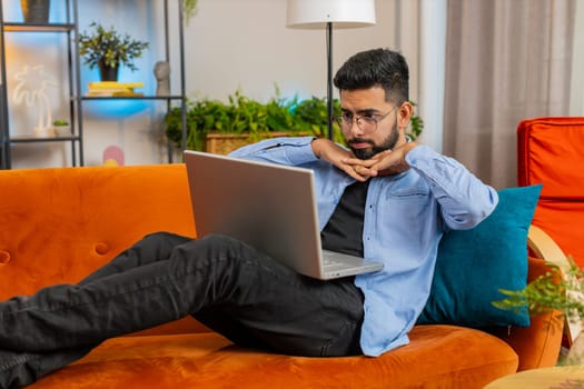 Upset young Indian man sitting on sofa at home using laptop having anxiety and stress. Shocked bearded Arabian guy reacting to loss, bad news on netbook in living room apartment. Computer virus.