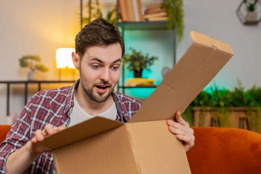Happy excited man unpacking delivery parcel sitting on sofa in living room. Smiling Caucasian guy shopper online shop customer showing thumbs up receiving purchase by fast postal shipping courier.