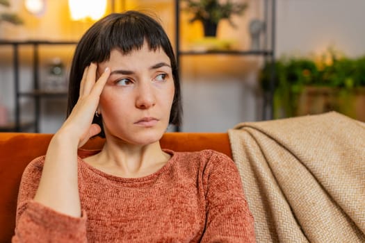 Sad lonely Hispanic girl looks pensive thinks over life concerns suffers from unfair situation sitting on sofa. Upset woman with hand in hair depressed feeling bad annoyed in living room at home.