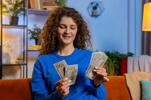 Successful happy rich woman with curly hair counting dollar banknotes while sitting on sofa in living room at home. Smiling girl calculating budget, savings, earnings, planning vacation in apartment.