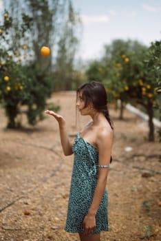 A young woman wearing a green dress is playfully tossing an orange in the air while standing in a picturesque orchard