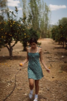A young woman walks through an orchard on a sunny day, wearing a green dress and holding an orange fruit.