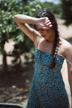 A young woman in a patterned dress stands outside in an orchard, shading her face from the sun with her hand