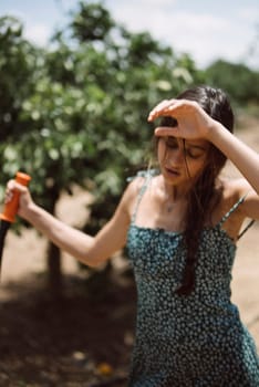 A young woman in a sundress outdoors on a sunny day, holding a drink and shielding her face from the sun.