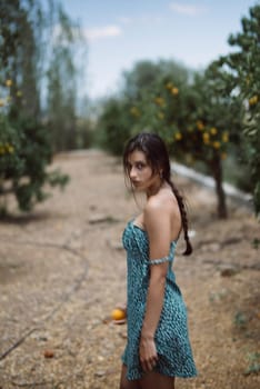 A young woman with braided hair stands in an orchard wearing a blue dress, surrounded by green trees.