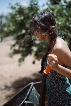 Side profile of a young woman holding an orange water gun, standing outdoors with lush greenery in the background