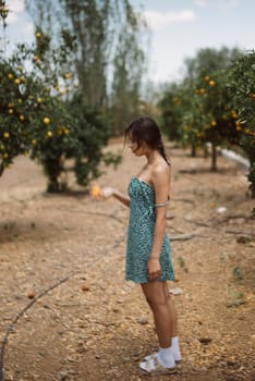 A woman in a green dress and sandals is standing in an orchard, holding an orange while surrounded by trees.