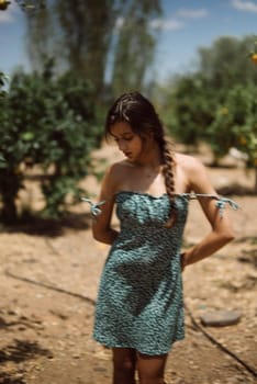 A young woman stands outdoors in a rural setting, wearing a summer dress with sunlight filtering through trees.