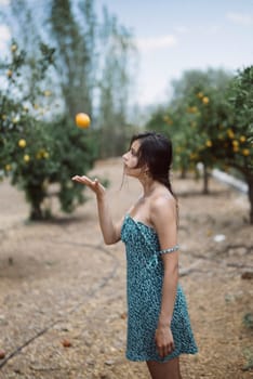 A woman wearing a blue dress is seen tossing an orange in a sunny orchard with trees in the background.