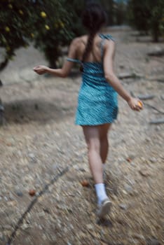 Young woman in blue dress walking through orchard, holding fruit, showcasing summer vibes and nature.