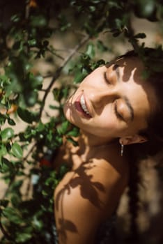 A woman is happily smiling with closed eyes, surrounded by lush greenery, and illuminated by sunlight