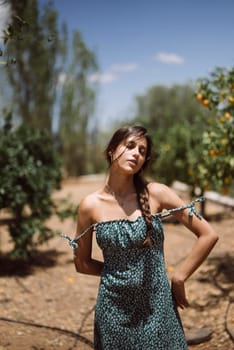 A woman posing outside in a summer dress with braided hair, surrounded by a green orchard on a sunny day