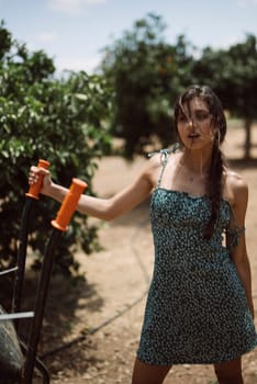 A woman is wearing a blue dress and holding garden tools among green foliage and trees on a sunny day