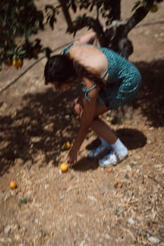 Woman bending down to pick fruit from the ground in a sunny orchard, wearing a green dress and white sandals.