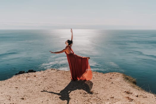 Woman red dress sea. Female dancer posing on a rocky outcrop high above the sea. Girl on the nature on blue sky background. Fashion photo