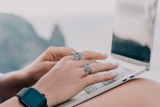 Digital nomad, woman in the hat, a business woman with a laptop sits on the rocks by the sea during sunset, makes a business transaction online from a distance. Freelance, remote work on vacation.