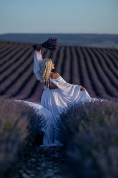 Blonde woman poses in lavender field at sunset. Happy woman in white dress holds lavender bouquet. Aromatherapy concept, lavender oil, photo session in lavender.