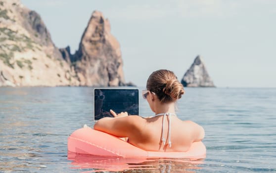 Woman freelancer works on laptop swimming in sea on pink inflatable ring. Pretty lady typing on computer while floating in the sea on inflatable donut at sunset. Freelance, remote work on vacation