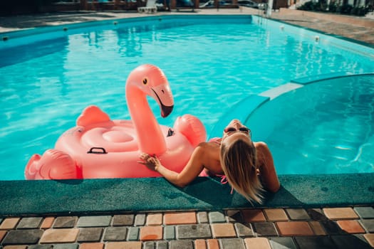 A woman in a pink bikini is sitting on a pink inflatable flamingo in a pool. The scene is playful and fun, with the woman enjoying her time in the water